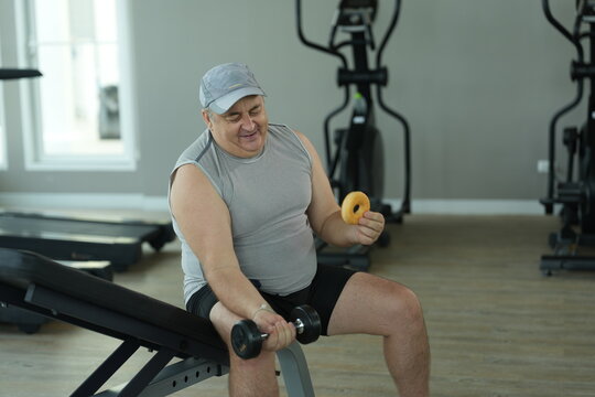 Overweight middle aged man in gray cap sits on the incline bench holding a dumbbell and a tempting donut during his weight loss workout at the fitness club in warm natural light