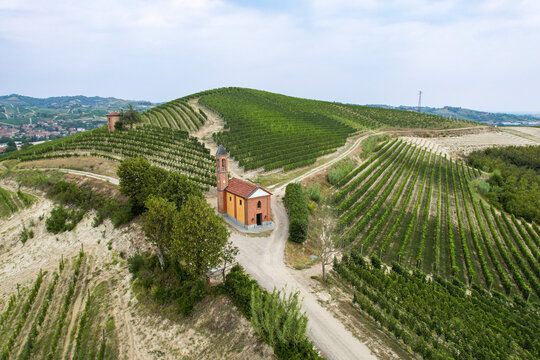 Aerial view of a small chapel nestled among lush green vineyard rows on rolling hills in Canale, Piedmont, Italy.