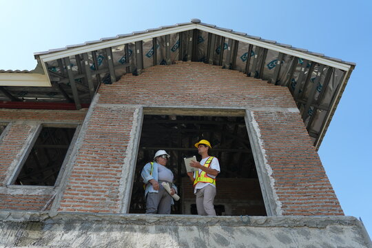 Two construction engineers in hard hats and safety vests review the specification sheets at the gable window at a large window opening of a new brick home being built in warm natural light