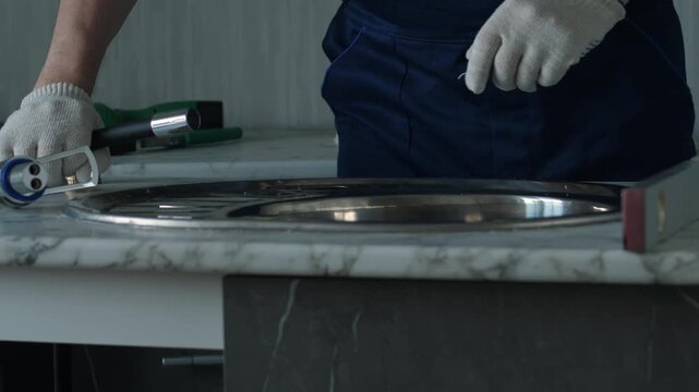 A plumber in protective gloves checks the horizontal alignment of a marble countertop with a spirit level. The professional worker then carefully fits a modern chrome faucet 