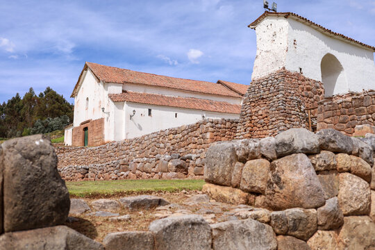 View of the historic Chinchero archaeological site featuring ancient Incan stone walls and a colonial church with a red tile roof under a blue sky in Cusco, Cusco, Peru.