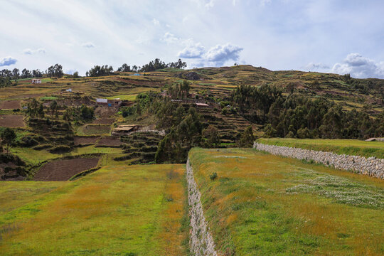 View of the ancient agricultural terraces and stone walls of Chinchero surrounded by rolling hills and trees under a cloudy sky in Cusco, Cusco, Peru.