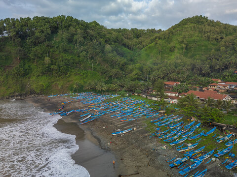 Ayah, Indonesia - 07 November 2022: Aerial view of Pasir Beach with numerous blue fishing boats lined up on the dark sand shore, surrounded by lush green hills and coastal vegetation.