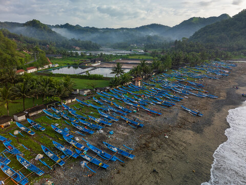 Ayah, Indonesia - 07 November 2022: Aerial view of Pasir Beach with blue fishing boats lined up on the sandy shore, surrounded by lush green hills and misty tropical landscape.