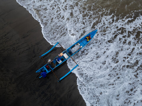 Ayah, Central Java, Indonesia - 07 November 2022: Aerial view of a traditional blue outrigger boat resting on the dark volcanic sand of Pasir Beach as white ocean waves wash ashore.