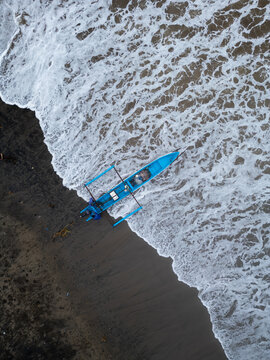 Ayah, Central Java, Indonesia - 07 November 2022: Aerial view of a blue outrigger boat on the dark volcanic sand of Pasir Beach as white ocean waves wash onto the shore.