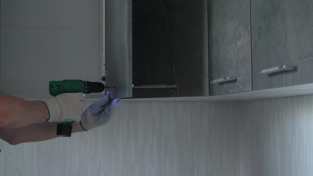 A professional craftsman in protective gloves uses an electric screwdriver to assemble kitchen cabinets. A handyman fastens screws into modern dark wooden furniture 