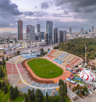 Ramat Gan, Israel - 04 March 2022: Aerial view of Ramat Gan Stadium with its green pitch and tiered seating, set against the modern skyline of Ramat Gan under a cloudy sky.