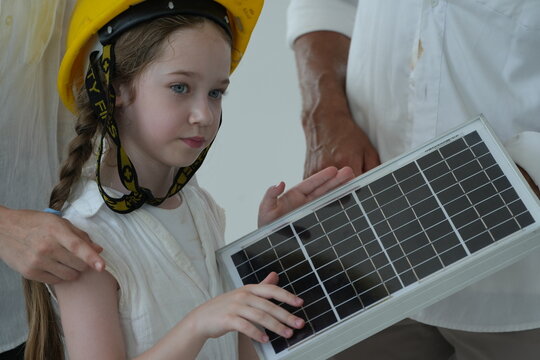 Happy young family with little daughter in a toy hard hat let the girl in a toy helmet touch the photovoltaic cells up close in their future eco friendly house on a bright sunny day