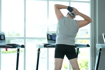 Fototapeta premium Overweight middle aged man in gray cap and sleeveless shirt does standing side stretches at the bright window front at the start of his new healthy lifestyle in the gym in warm natural light