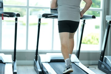 Fototapeta premium Funny fat senior man in sportswear walks the treadmill in close up, step after heavy step at the start of his new healthy lifestyle in the gym on a bright sunny day