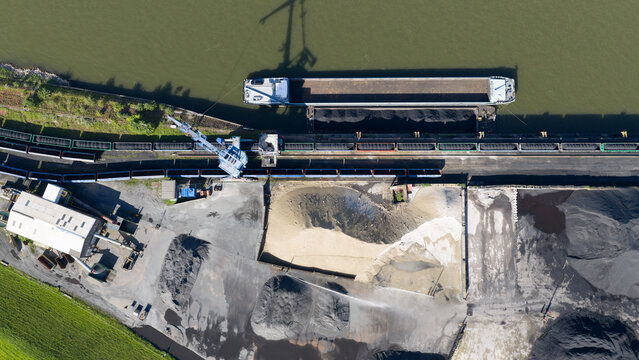 Aerial view of an industrial port facility featuring a barge loaded with bulk materials, a large crane, and stockpiles of coal and sand in Duisburg, North Rhine-Westphalia, Germany.