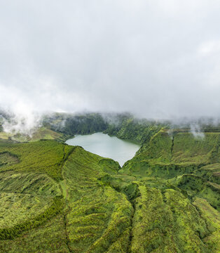 Aerial view of the volcanic crater lake at Miradouro Caldeira Rasa e Funda surrounded by lush green hills and mist in Mosteiro, Acores, Portugal.