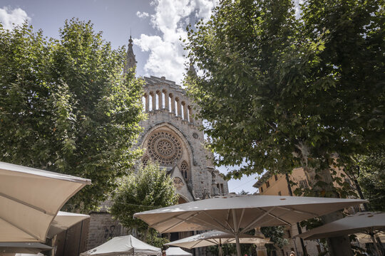 View of the Church of Saint Bartholomew with its ornate rose window framed by lush green trees and outdoor cafe umbrellas in the town square of Soller, Balearic Islands, Spain.