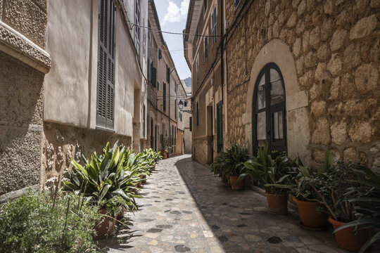 View of a narrow cobblestone street lined with traditional stone buildings and lush potted plants under bright sunlight in Soller, Balearic Islands, Spain.