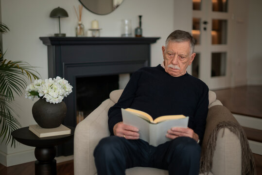 Grey haired senior man sitting in an armchair and reading book in domestic room