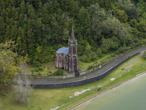 Aerial view of Ermida de Nossa Senhora das Vitorias, a Gothic revival chapel nestled in lush forest beside the lake with a stone wall and path in Furnas, Acores, Portugal.