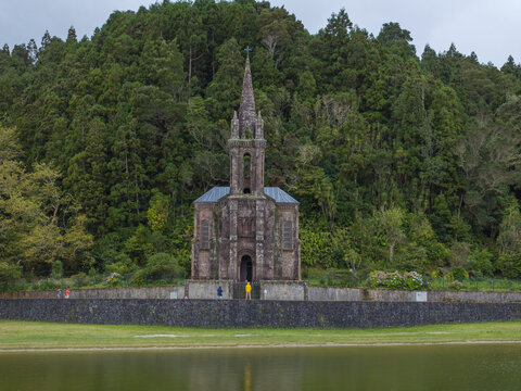 Aerial view of Ermida de Nossa Senhora das Vitorias, a historic stone chapel nestled by a lake and surrounded by dense forest with people standing nearby in Furnas, Acores, Portugal.