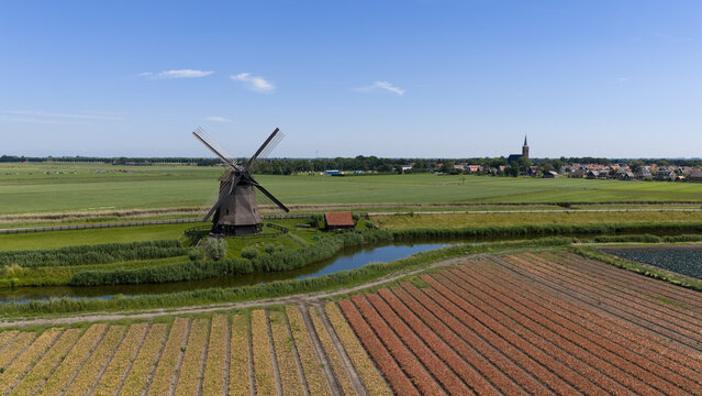 Aerial view of Bovenmolen G windmill surrounded by lush green fields, a winding canal, and agricultural rows under a clear blue sky in Schermerhorn, Noord-Holland, Netherlands.