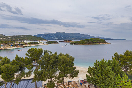 Aerial view of the turquoise waters and small islands of Ksamil, with lush pine trees and a sandy beach under a cloudy sky in Ksamil, Vlore County, Albania.