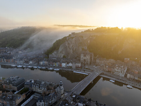 Aerial view of the Collegiate Church of Notre-Dame de Dinant and the Citadel perched on the cliff above the Meuse River during a misty golden hour in Dinant, Wallonia, Belgium.