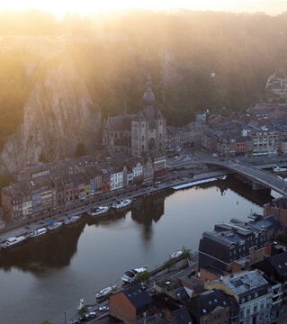 Aerial view of the Collegiate Church of Notre Dame de Dinant nestled against a dramatic limestone cliff along the Meuse River at golden hour in Dinant, Wallonia, Belgium.