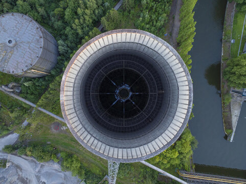 Aerial view of the massive circular cooling tower of the abandoned IM power plant, surrounded by lush green trees, a river, and industrial structures in Charleroi, Wallonia, Belgium.