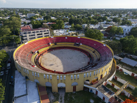 Aerial view of Plaza De Toros Merida, a historic bullring with vibrant red and yellow seating, surrounded by lush trees and urban buildings in Merida, Yucatan, Mexico.