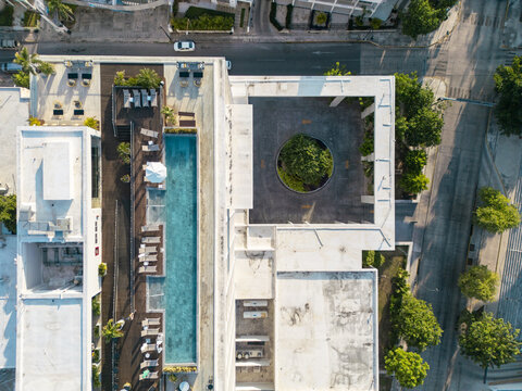 Aerial view of a modern hotel building featuring a long rectangular swimming pool, sun loungers, and a circular courtyard with a central tree in Merida, Yucatan, Mexico.