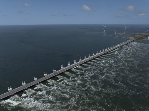 Aerial view of the Eastern Scheldt Storm Surge Barrier with turbulent water currents, wind turbines in the distance, and a clear blue sky in Vrouwenpolder, Zeeland, Netherlands.