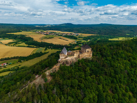 Aerial view of Tocnik Castle perched on a forested hill, surrounded by rolling agricultural fields and meadows under a cloudy blue sky in Tocnik, Stredocesky kraj, Czechia.