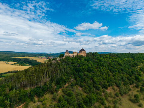 Aerial view of Tocnik Castle situated on a forested hill surrounded by rolling fields and a blue sky with scattered clouds in Tocnik, Stredocesky kraj, Czechia.