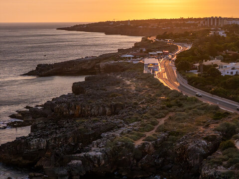 Aerial view of Boca do Inferno at sunset with rugged cliffs, coastal road, and the Atlantic Ocean in Cascais, Lisboa, Portugal.