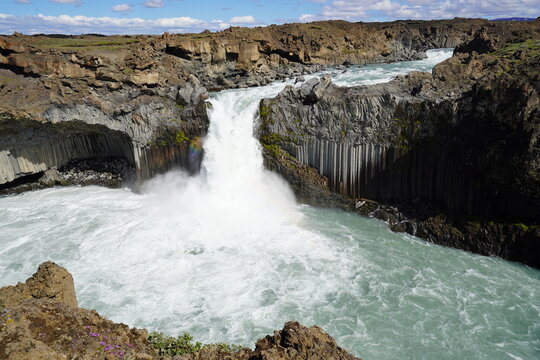 View of Aldeyjarfoss waterfall cascading over basalt columns into a turquoise pool surrounded by rugged volcanic rock formations under a bright blue sky in Iceland.