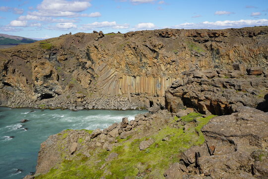 View of the dramatic basalt column rock formations along the river canyon with mossy green slopes under a blue sky in Thingeyri, Isafjardarbaer, Iceland.