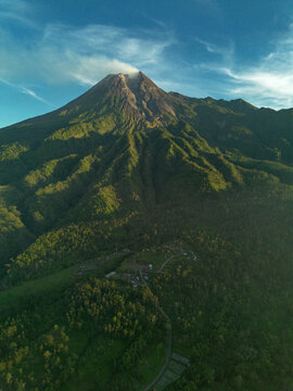 Aerial view of Mount Merapi volcano with smoke rising from the crater, surrounded by lush green forested slopes and small village settlements at the base in Indonesia.