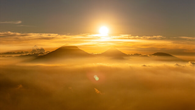 Aerial view of the volcanic peaks of Mount Sumbing and Mount Sindoro rising above a dense sea of clouds during a golden sunset in Selo, Central Java, Indonesia.