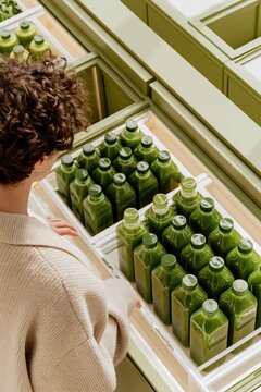 Curly haired woman choosing green juice from a refrigerated display case in a grocery store, fresh bottled beverages in a bright retail setting for healthy lifestyle content