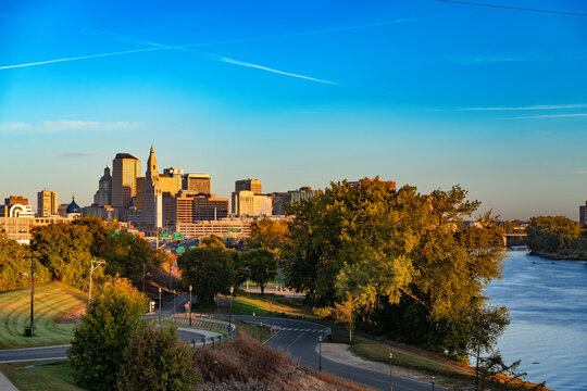 A panoramic view of downtown Hartford, Connecticut at sunrise showing city skyscrapers, lush trees along Charter Oak Landing park, and the calm waters of the Connecticut River.