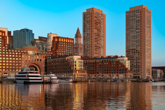 Panoramic view of downtown Boston skyline and the waterfront as seen from the Seaport district, featuring Rowes Wharf, tall residential towers, boats docked in the harbor, and golden sunset light.