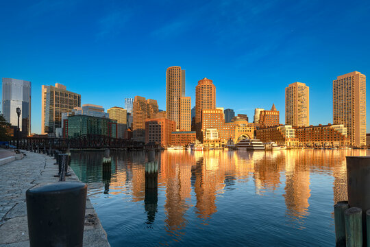 Golden morning light at sunrise over the downtown Boston cityscape with reflections on the calm harbor water, as viewed across the bay from the Seaport District in the early morning.