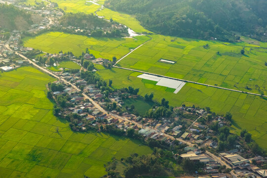 Aerial view of Lak Lake surrounded by lush green rice paddies and a small village with houses along a road in Lien Son Lak, Dak Lak, Vietnam.