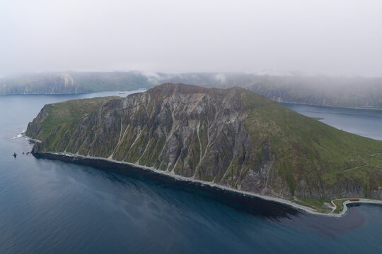 Aerial view of Mount Ballyhoo with rugged green slopes, rocky cliffs, and dark volcanic sand beaches surrounded by the deep blue waters of the Bering Sea in Unalaska, Alaska, United States.