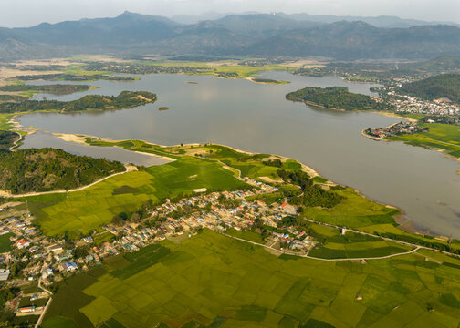 Aerial view of Lak Lake surrounded by vibrant green rice paddies and a small village with mountains in the distance in Lien Son Lak, Dak Lak, Vietnam.