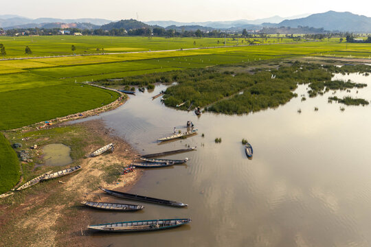 Aerial view of Lak Lake with wooden boats moored along the muddy shore, surrounded by vibrant green rice paddies and distant mountains in Lien Son Lak, Dak Lak, Vietnam.