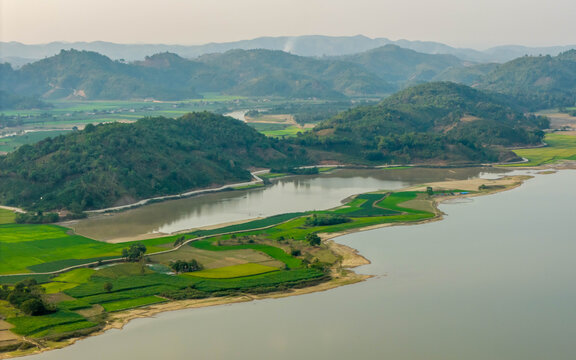 Aerial view of Lak Lake surrounded by lush green rice paddies and rolling hills under a soft sky in Lien Son Lak, Dak Lak, Vietnam.
