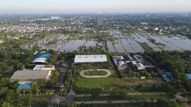 Aerial view of Jakabaring sports complex with a running track and large buildings surrounded by flooded fields and urban landscape in Kecamatan Seberang Ulu I, Sumatera Selatan, Indonesia.