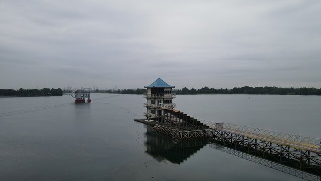 Aerial view of the Jakabaring Sport City lake featuring a building with a blue roof and wooden pier, surrounded by calm water in Kecamatan Seberang Ulu I, Sumatera Selatan, Indonesia.