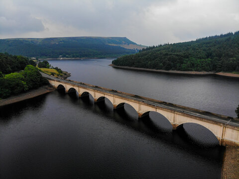 Aerial view of Ladybower Reservoir with its iconic stone arched bridge spanning the dark water, surrounded by lush green hills under an overcast sky in Hope Valley, England, United Kingdom.