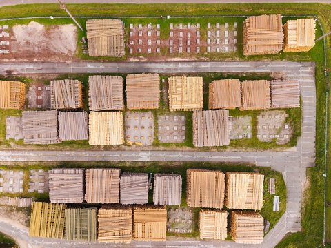 Aerial view of stacked lumber and timber piles arranged in an industrial yard with concrete foundations and surrounding grass in Brankas, Jelgava Municipality, Latvia.